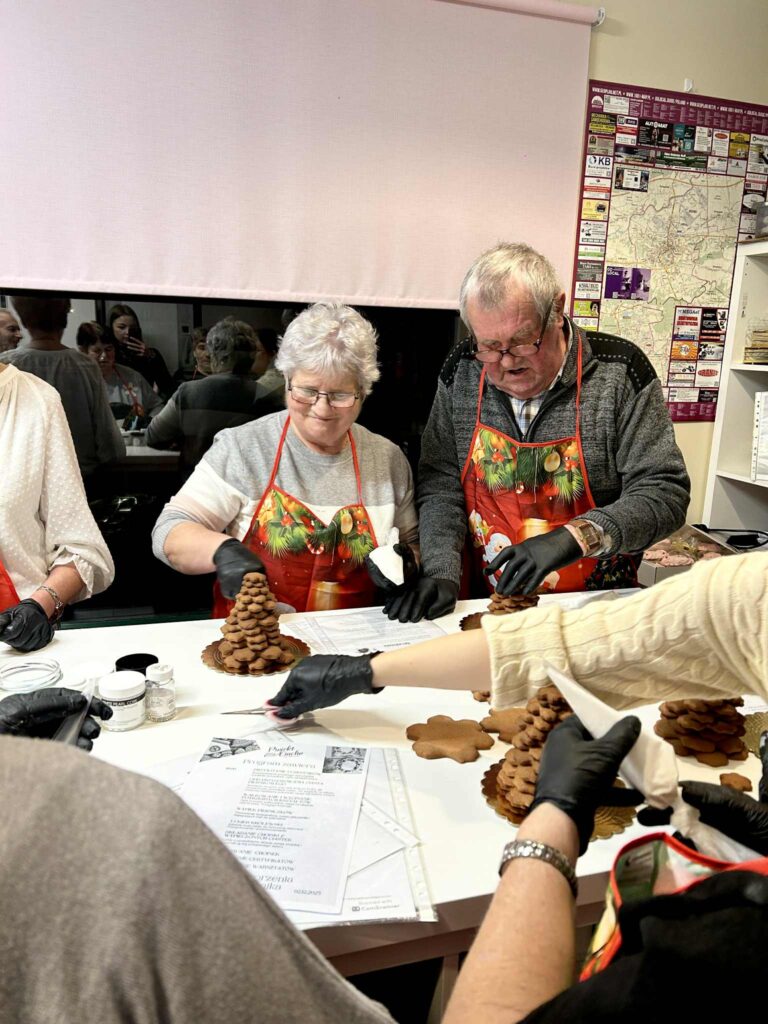 Zdjecie przedstawiające uczestników warsztatów - małżeństwo seniorów, którzy układaja z pierników choinkę